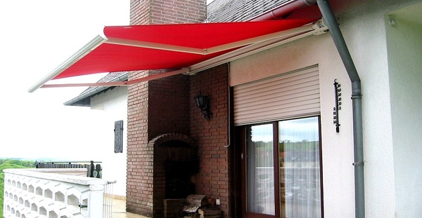 Rote Markise als Sonnenschutz über Terrasse an Haus mit Fenster und gemauertem Grill.